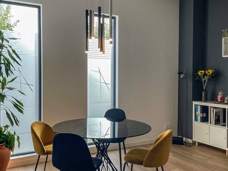 Detalle de zona de reuniones con mesa reconda de cristal y sillas tipo butaca tapizadas en color mostaza y negro. Cristalera vertical al fondo con vinilo al ácido. Vista exterior a la calle principal. Suelo de cerámica imitación madera. Foto Ángela Fernández