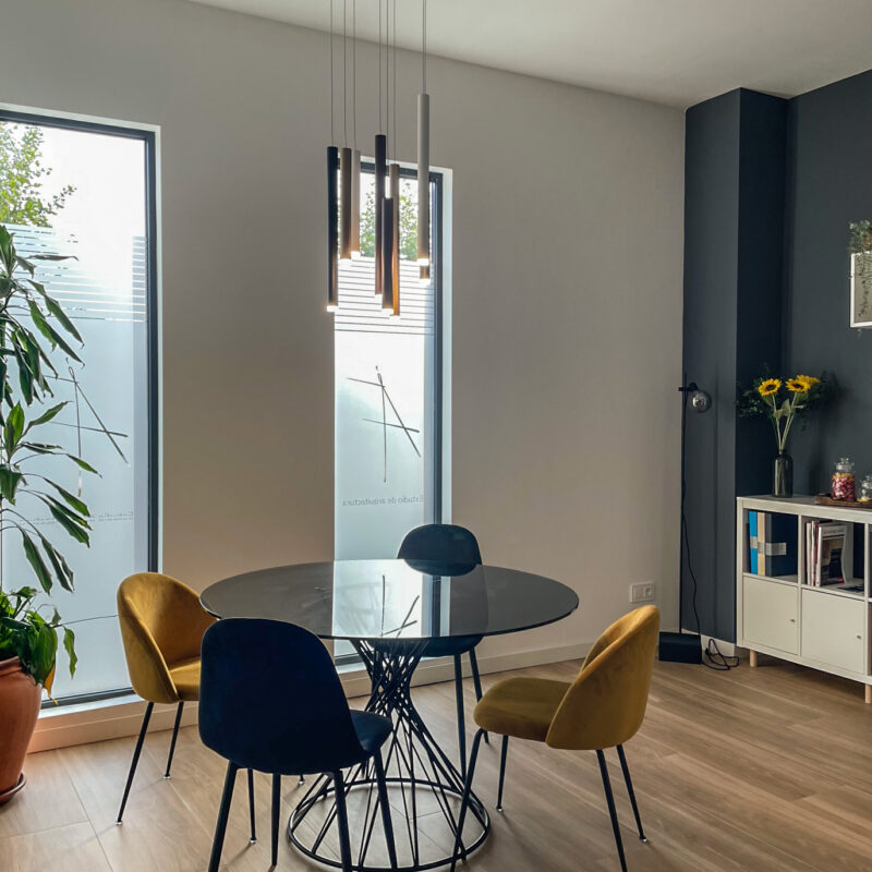 Detalle de zona de reuniones con mesa reconda de cristal y sillas tipo butaca tapizadas en color mostaza y negro. Cristalera vertical al fondo con vinilo al ácido. Vista exterior a la calle principal. Suelo de cerámica imitación madera. Foto Ángela Fernández